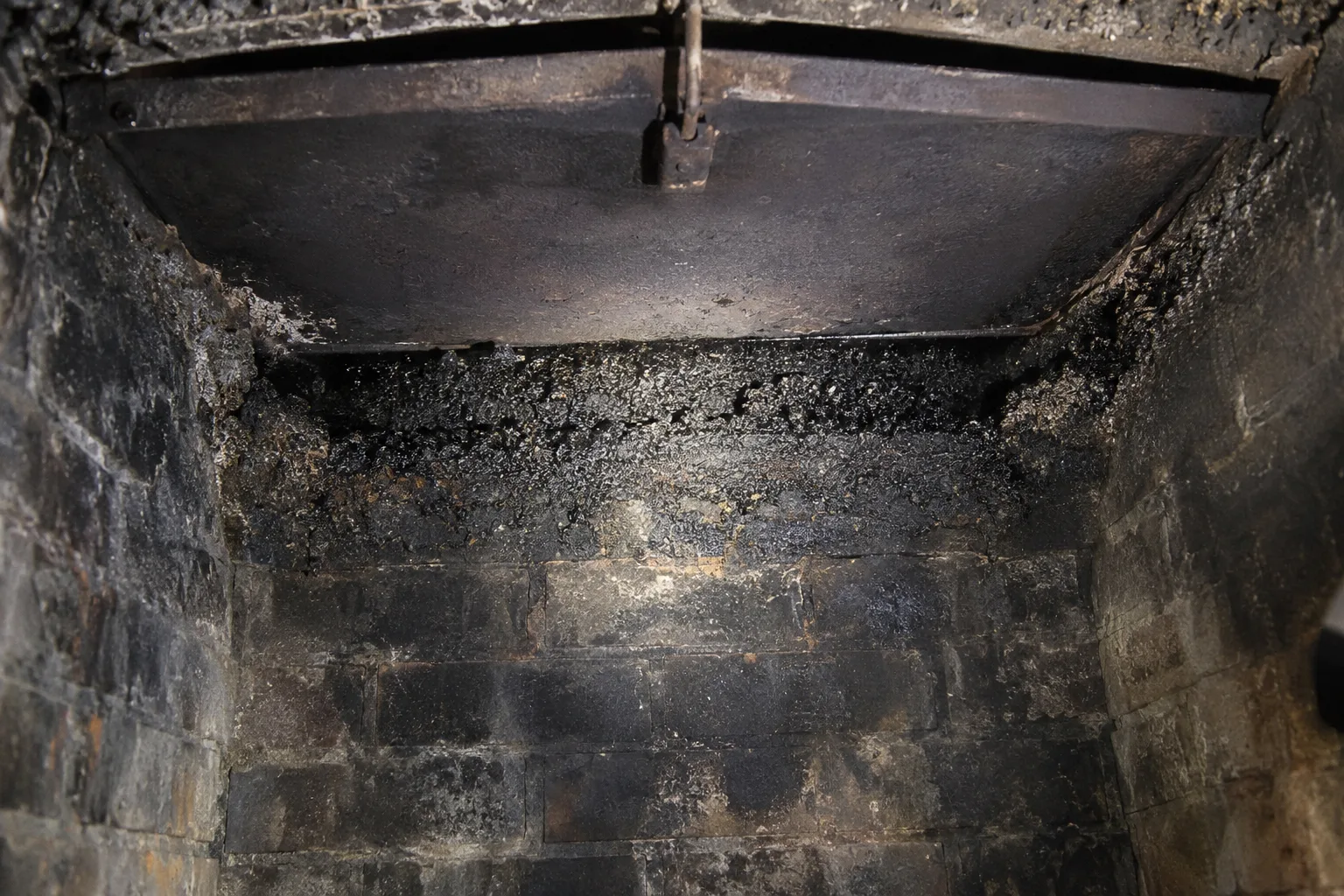 View looking up inside a masonry fireplace firebox at the smoke shelf and damper, both coated in thick black soot and hardened creosote deposits, lit by a work light