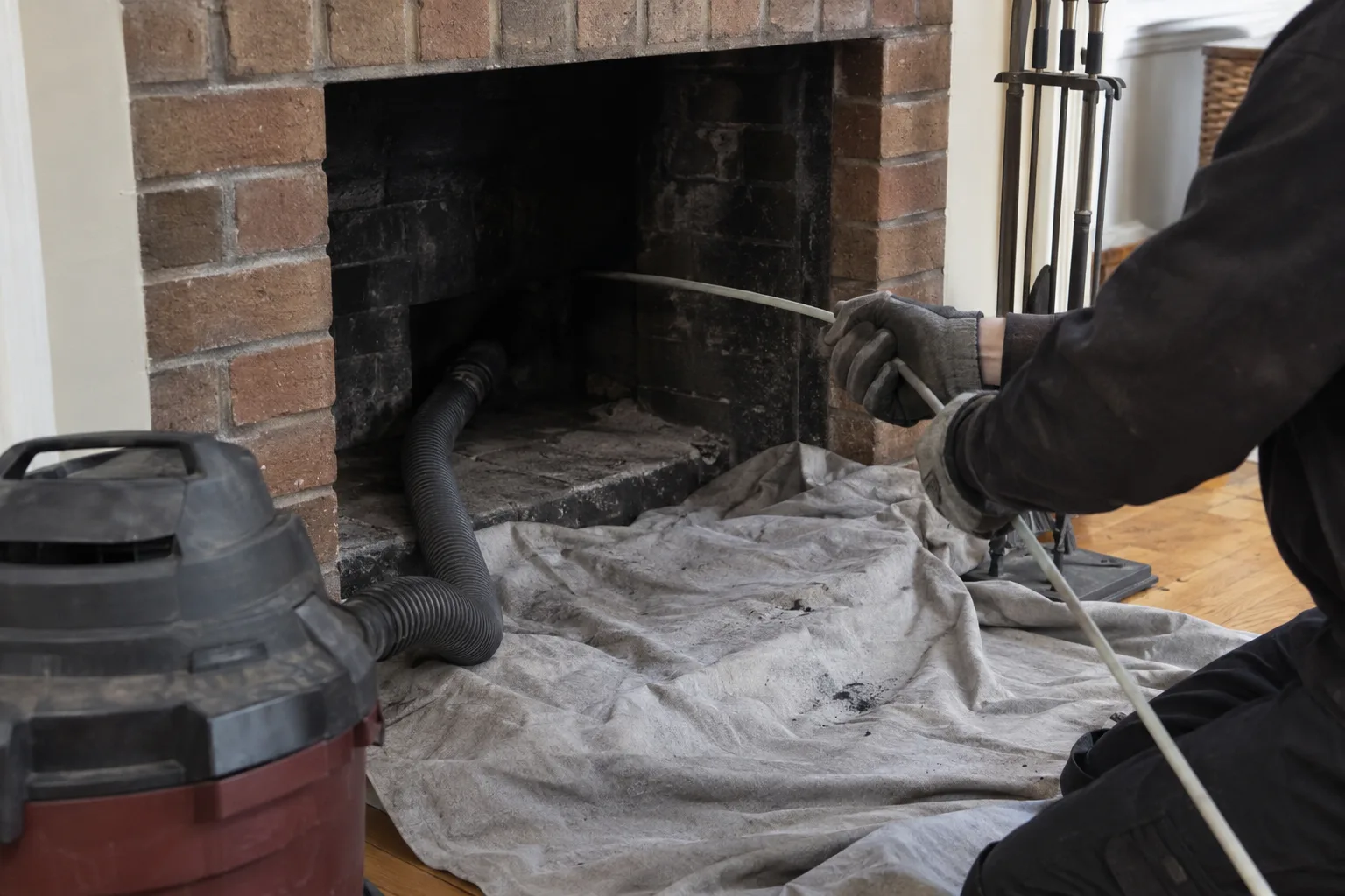 Gloved hands feeding a flexible cleaning rod into the opening of a brick firebox, with a drop cloth protecting the hearth floor and a shop vacuum connected nearby