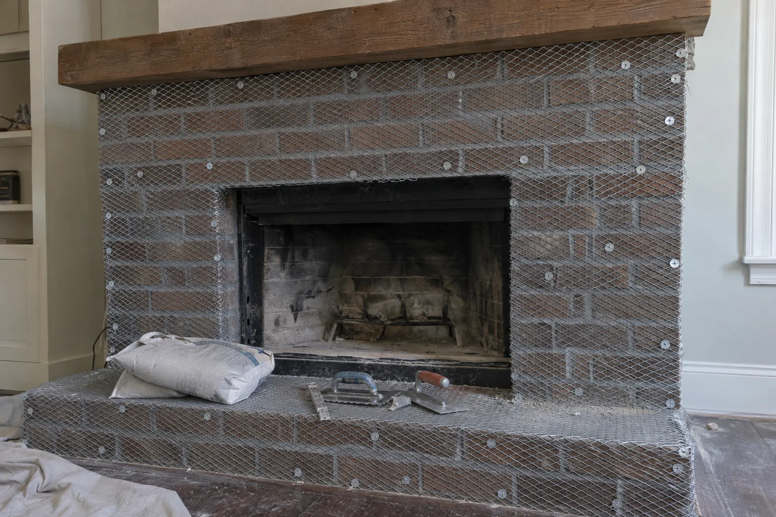 Galvanized metal lath mesh fastened across an entire brick fireplace surround as a bonding substrate layer, with a bag of modified thinset and masonry tools staged on the hearth below