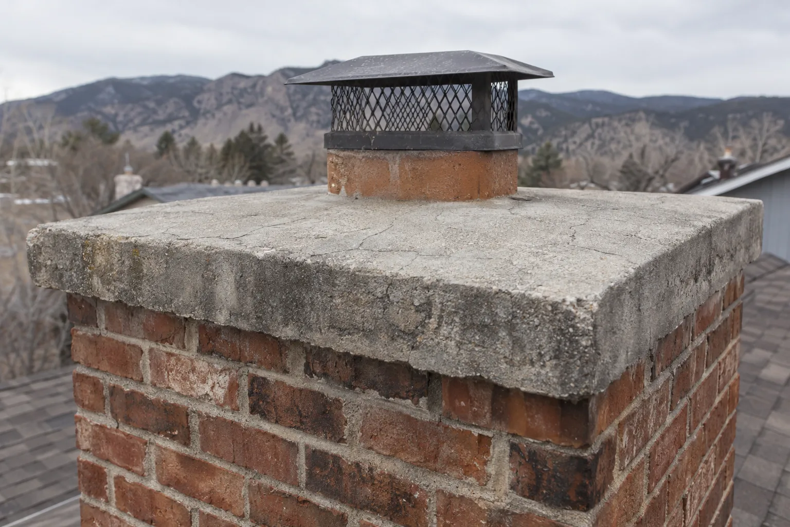 Weathered concrete chimney crown with a pattern of fine crazing cracks across its surface and a two-inch overhang edge, with blurred roof shingles and Colorado foothills in the background