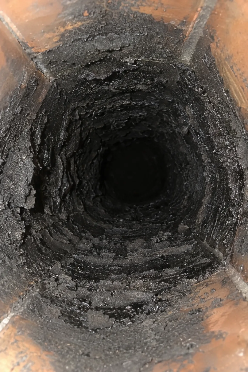 View looking up inside a clay tile chimney flue showing heavy black creosote deposits coating the interior walls, illuminated by a work light from below
