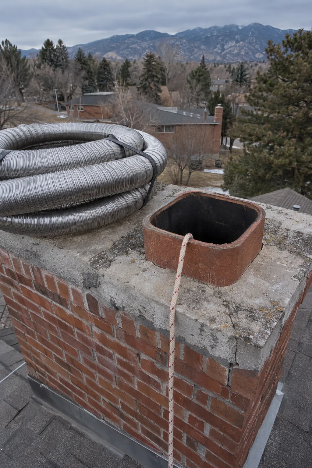 Rooftop of a brick home with a masonry chimney in the foreground, a coiled corrugated stainless steel flexible liner resting on the chimney crown beside the open flue, a guide rope already threaded into the flue opening under soft Colorado morning light