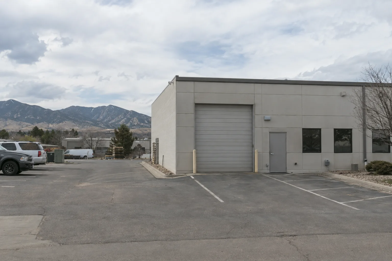 A small light industrial unit in a Colorado business park with a roll-up door and painted parking spaces in front, the Rocky Mountain foothills visible in the distance under a partly cloudy sky