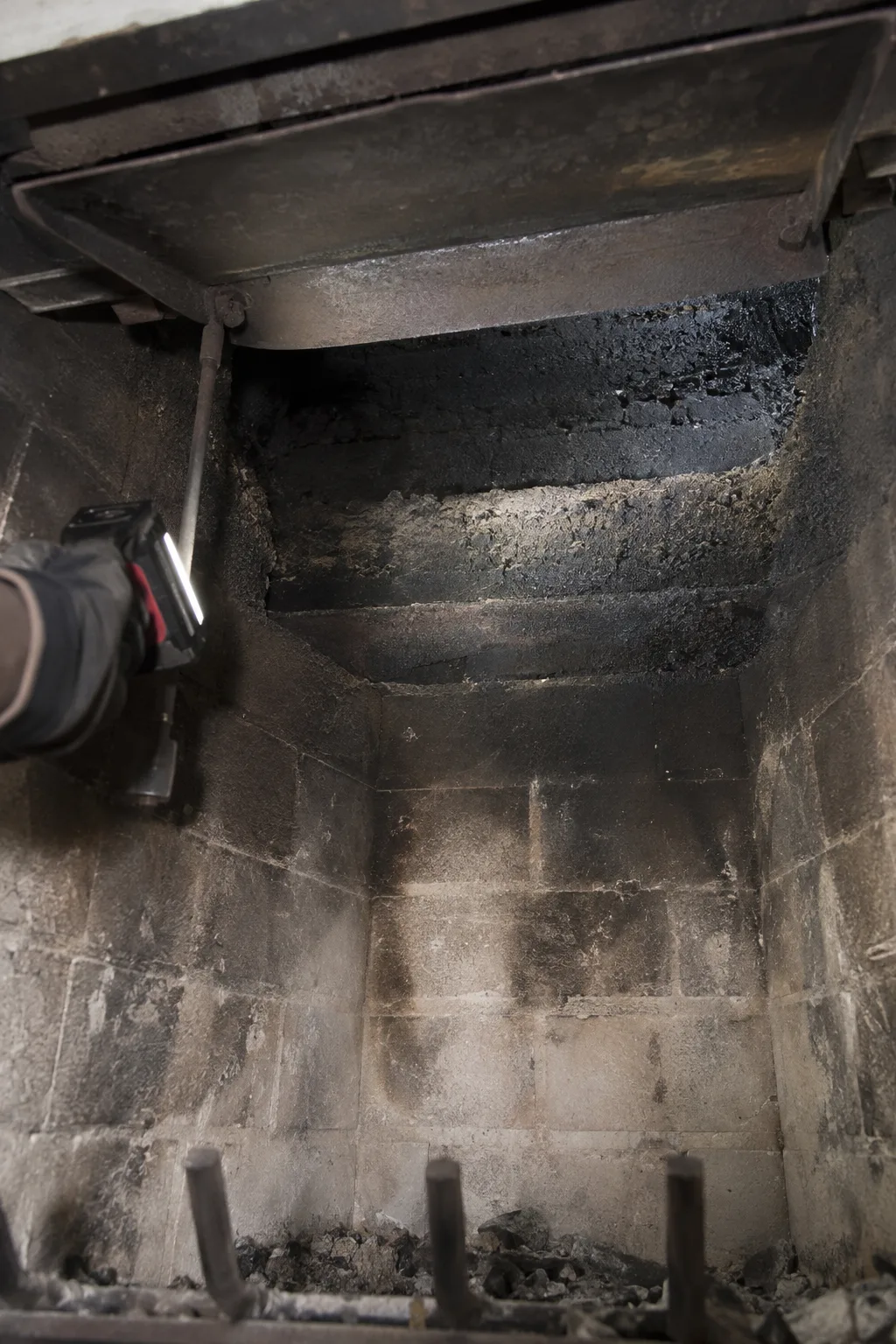 View from inside a masonry firebox looking upward past an open cast-iron damper plate toward a heavily sooted smoke shelf, lit by a handheld LED inspection light held by a gloved hand at the edge of the frame