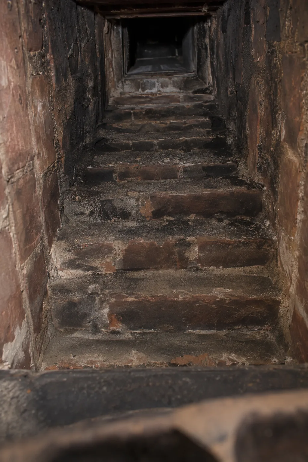Upward view from inside a fireplace through the open damper throat into a corbeled brick smoke chamber, showing stair-stepped ledges with dark creosote deposits in the corners and a narrow rectangular flue opening at the top, illuminated by a flashlight beam