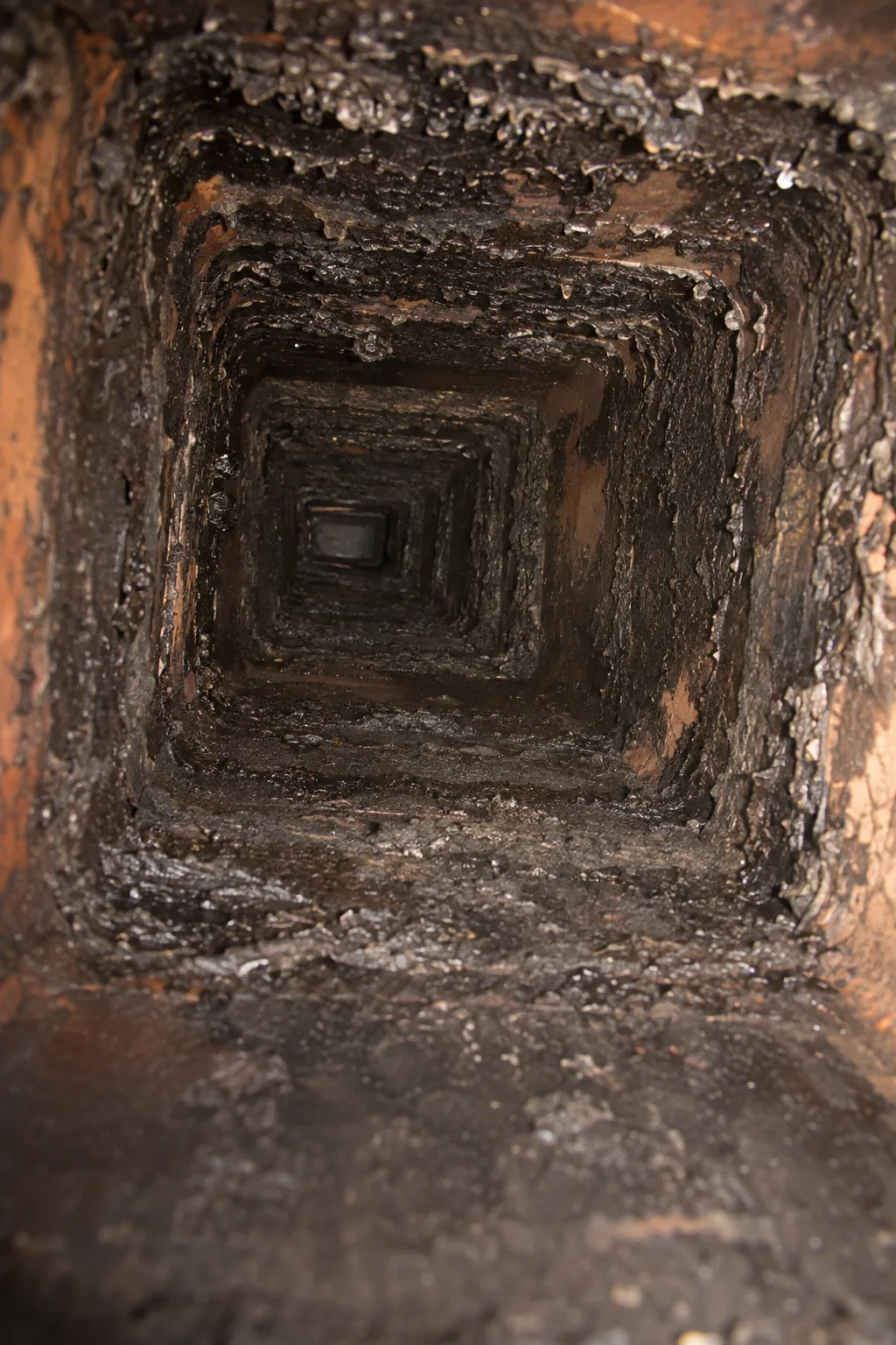 View looking up the inside of a rectangular clay chimney flue liner whose walls are coated with dark, flaky creosote deposits, lit by a handheld work light