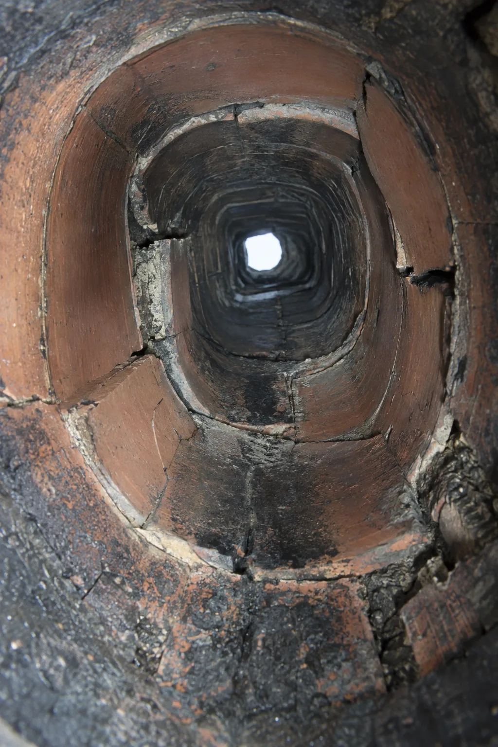 Upward-looking interior camera view of a deteriorated chimney flue showing cracked and offset terra cotta tile liner sections with visible fractures, spalling edges, and dark creosote staining illuminated by a bright camera light