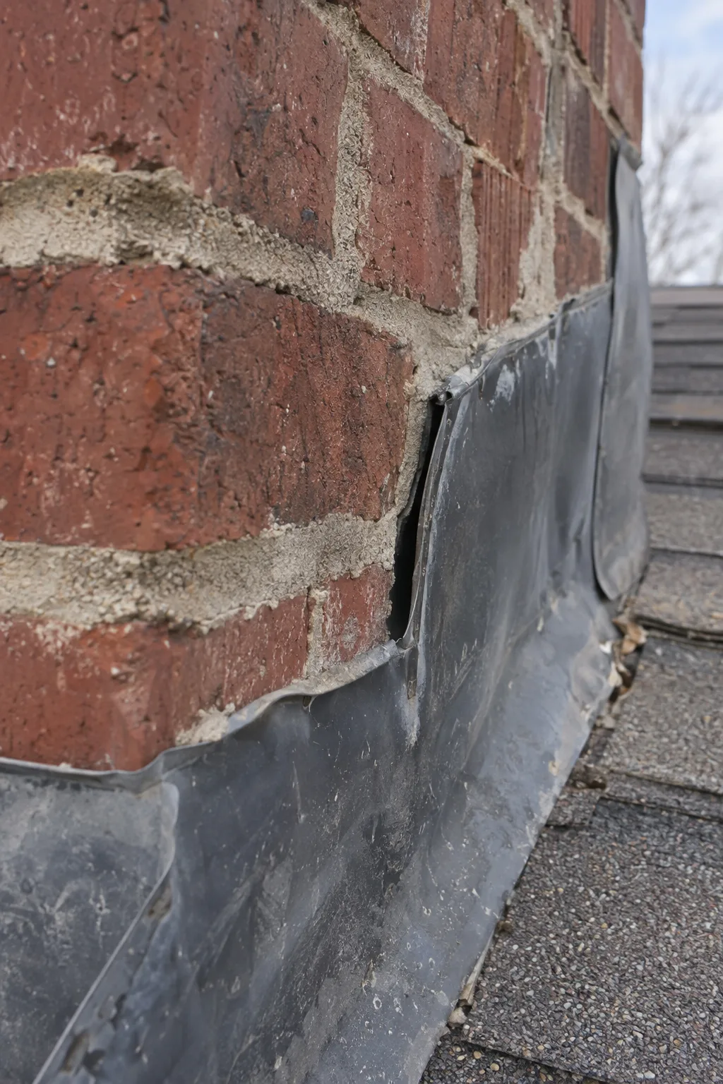 Close-up of a brick chimney base showing metal counterflashing separated from the mortar joint, with a visible gap between the bent metal lip and the masonry surface